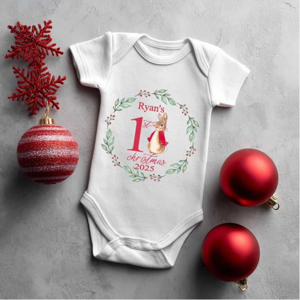 White baby onesie with 'Ryan's 1st Christmas 2025' design, surrounded by red Christmas ornaments on a gray surface.