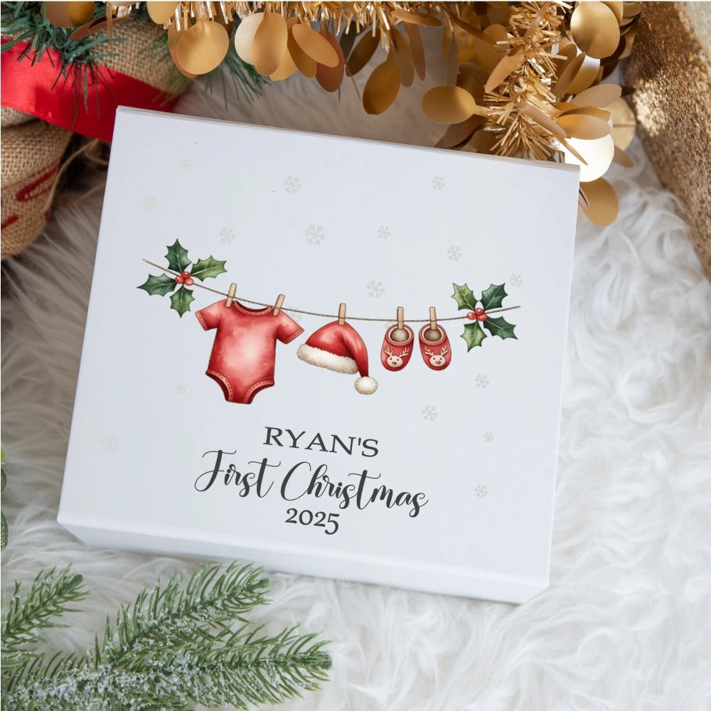Personalised baby Christmas keepsake box showing a festive clothesline with a red baby bodysuit, Santa hat and reindeer slippers hanging between holly leaves; the lid reads “Ryan’s First Christmas 2025”.
