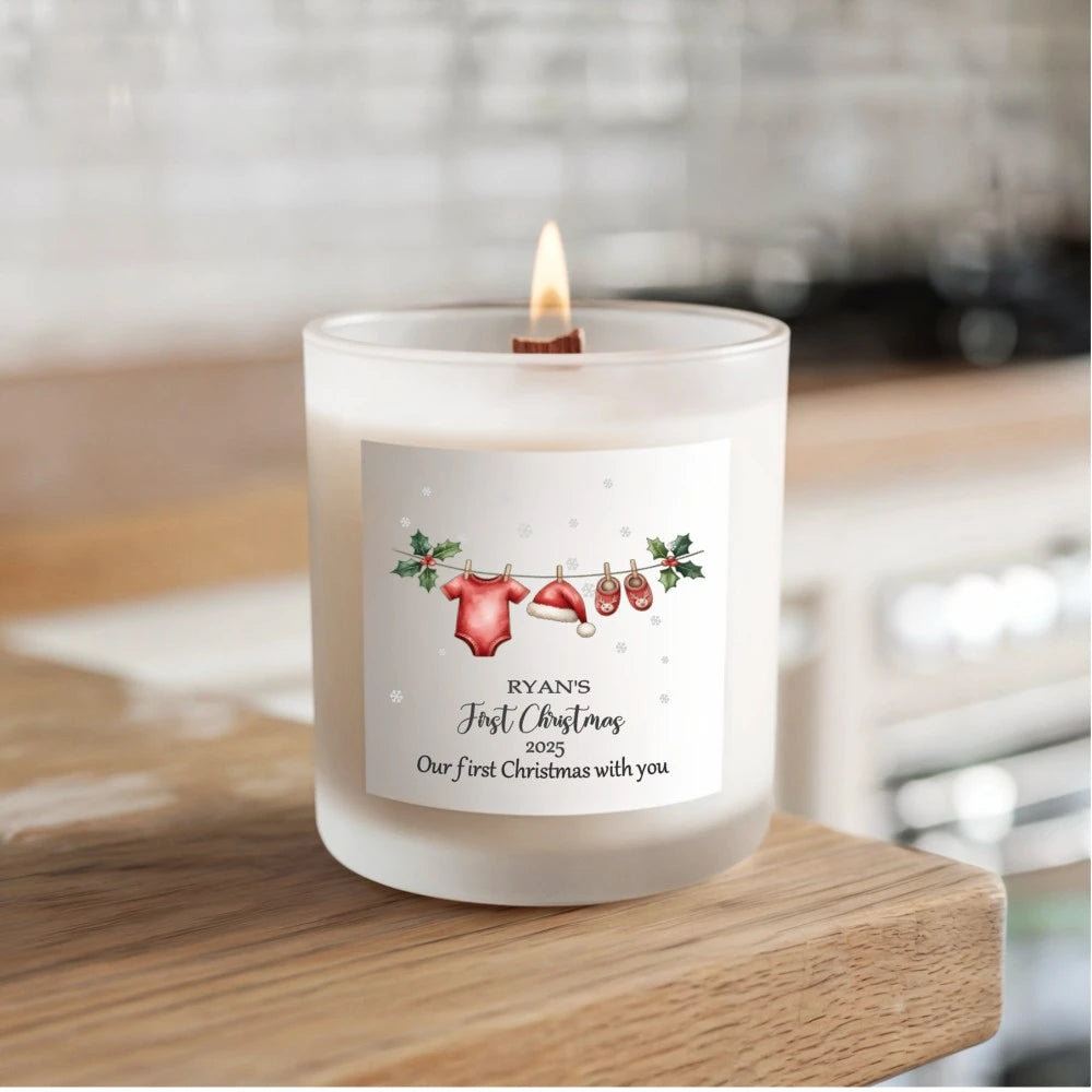 Personalised soy candle showing a festive clothesline with a red baby bodysuit, Santa hat and reindeer slippers hanging between holly leaves, with baby's name and First Christmas 2025
