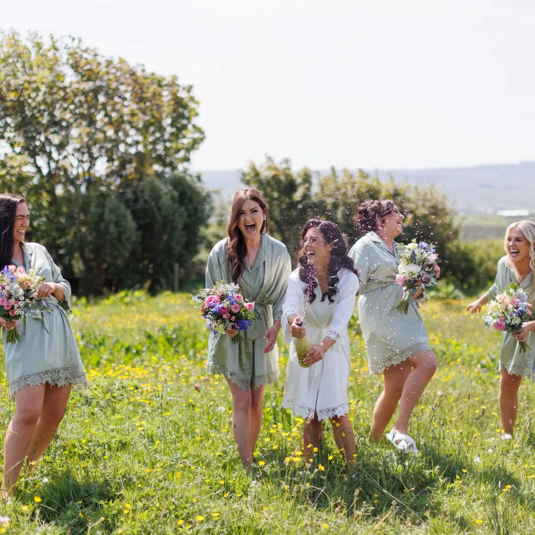 Bride and bridesmaids wearing matching personalised satin wedding robes in sage green and white, celebrating outdoors in Ireland with bouquets and champagne — handmade by HanaLee Studios, Co. Meath