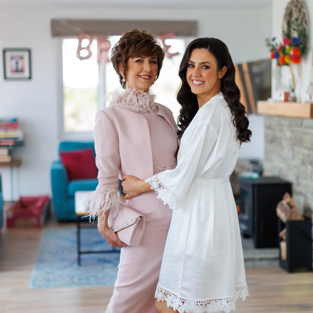Bride in personalised white lace-trim satin robe standing with mother of the bride in blush outfit on wedding morning — Irish handmade wedding robe by HanaLee Studios.