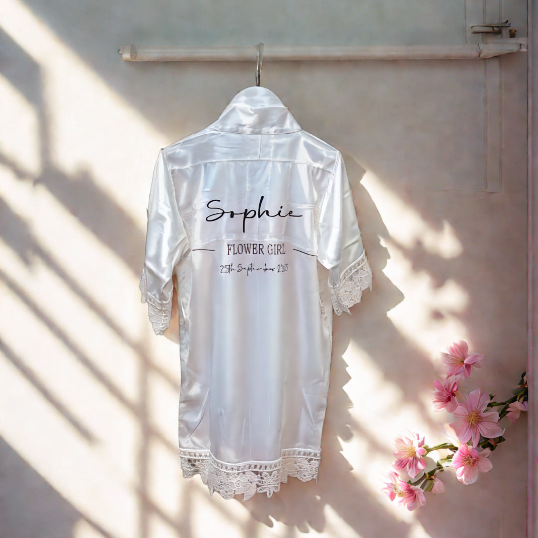 Personalised satin flowergirl robe in white with lace trim, customised with name ‘Sophie’ and wedding date, handmade in Ireland by HanaLee Studios.