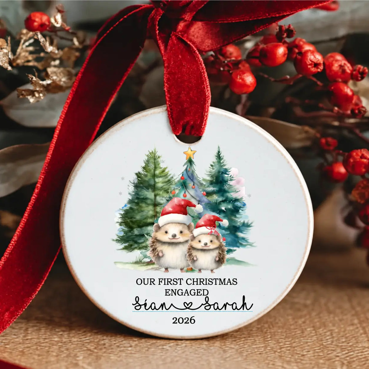 Personalised Our First Christmas Engaged 2026 ceramic Christmas decoration featuring two cute hedgehogs in Santa hats, Christmas trees and couple's names, finished with a red velvet ribbon.