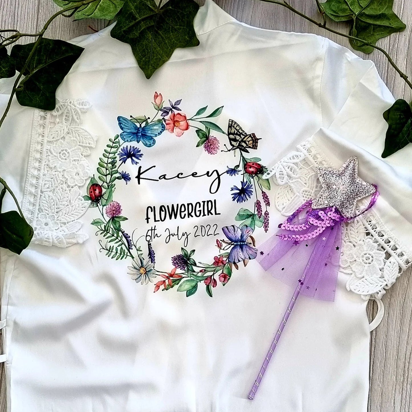 Personalised white satin flowergirl robe with lace trim and wildflower wreath design, customised with name and date — designed and finished in Ireland by HanaLee Studios.