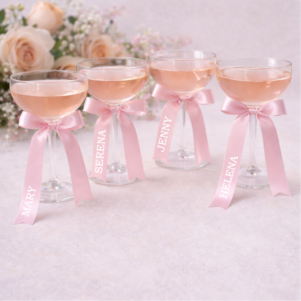 Four cocktail glasses styled with pale pink satin wedding ribbons tied in bows, personalised with names, photographed on a soft neutral background with blush flowers