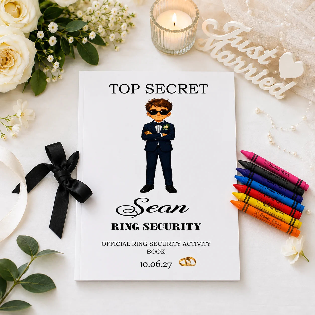 Personalised Ring Security activity book for page boys featuring a ring security character, child's name and wedding date, displayed with crayons on a wedding-themed table setting with black ribbon and floral decorations.