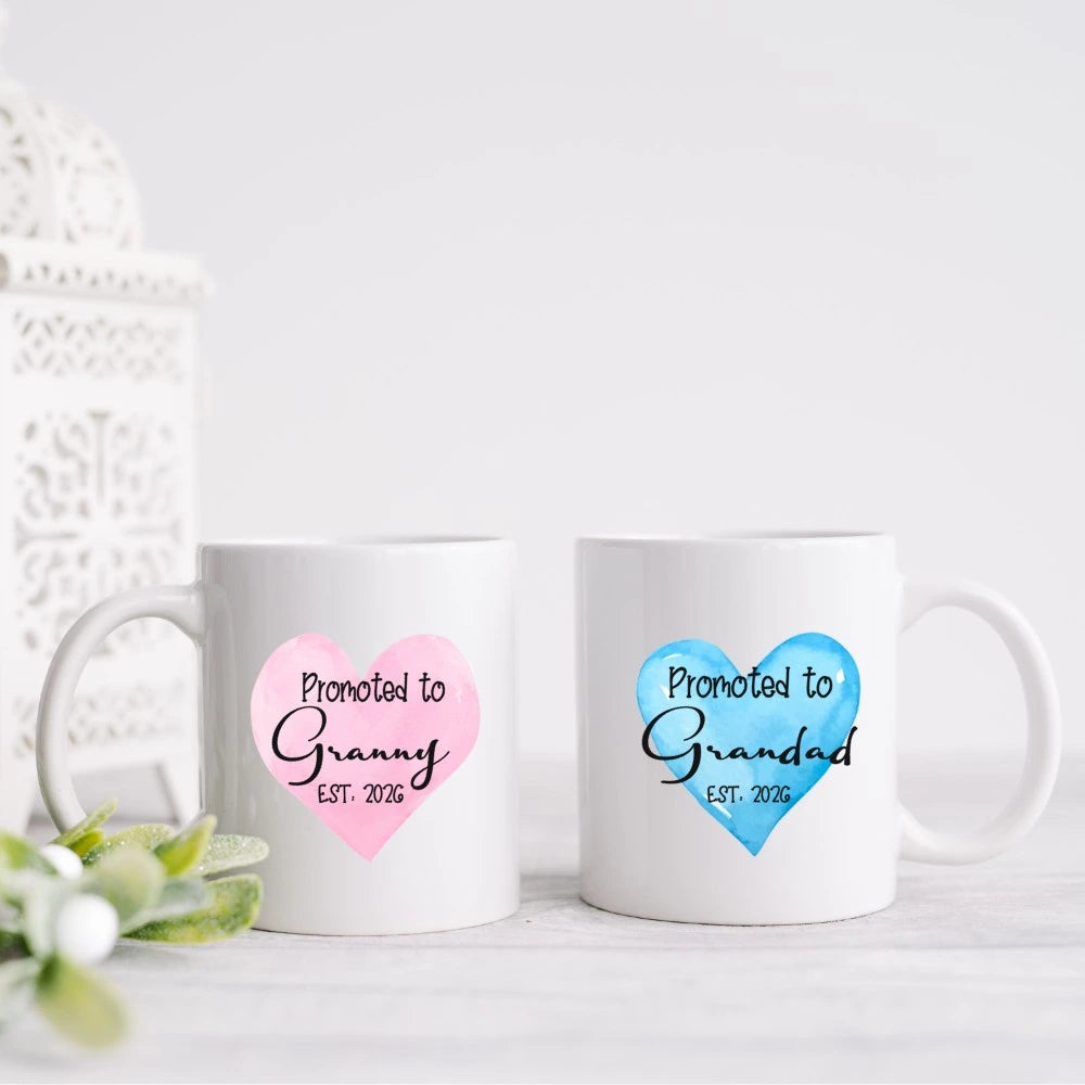 White ceramic mugs with pink and blue heart designs reading Promoted to Granny and Promoted to Grandad, personalised pregnancy announcement gift handmade in Ireland.
