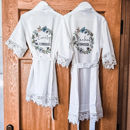 Two flowergirl robes hanging on a door in white fabric with butterfly wreath design and personalised with name, title and date.