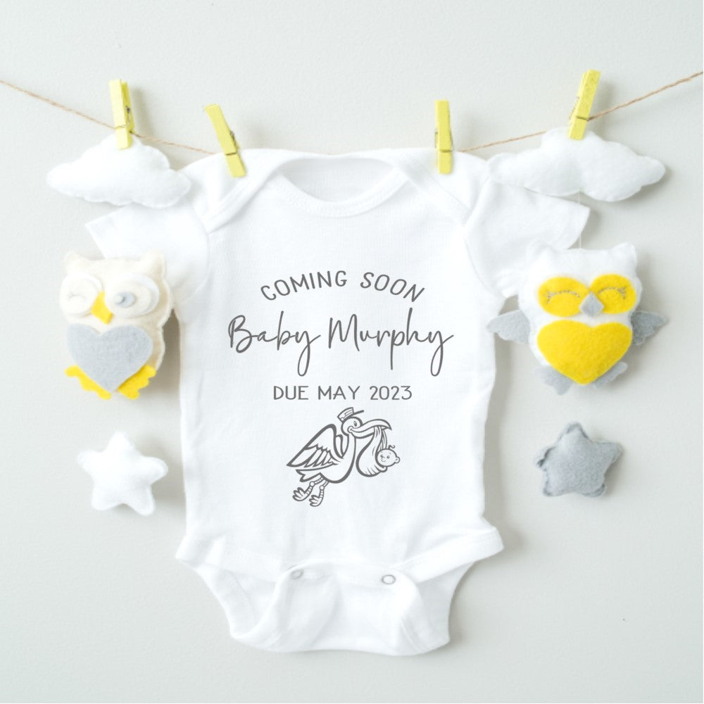 A white baby announcement vest with text and illustration, displayed on a clothesline with decorative elements around.