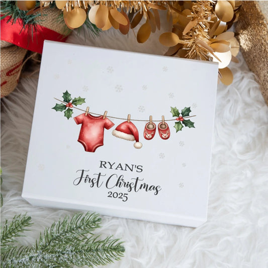 Personalised baby Christmas keepsake box showing a festive clothesline with a red baby bodysuit, Santa hat and reindeer slippers hanging between holly leaves; the lid reads “Ryan’s First Christmas 2025”.