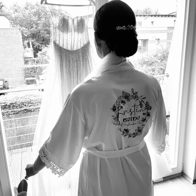 Bride wearing a personalised satin robe printed with floral wreath design, standing by a window admiring her wedding dress, robe printed by HanaLee Studios Ireland.