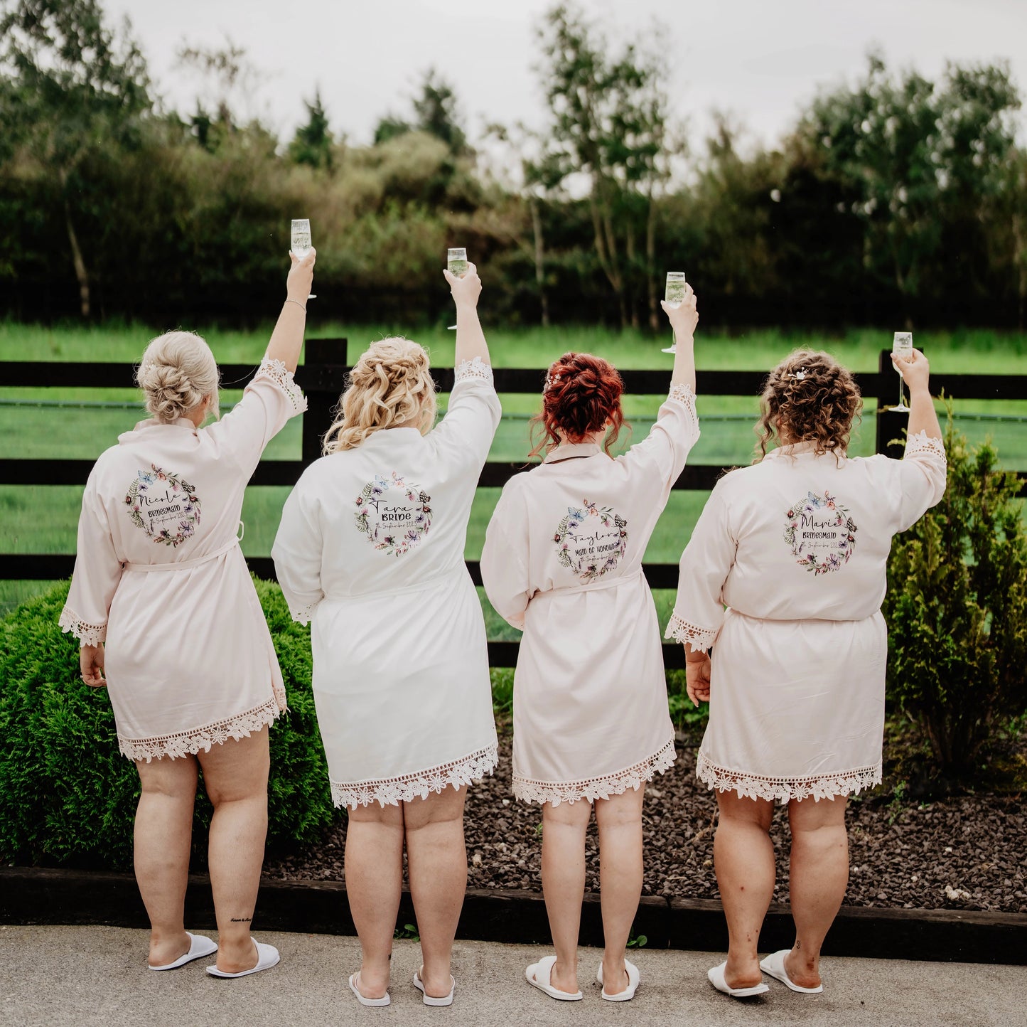 Bride and bridesmaids wearing matching personalised satin robes with lace trim, raising champagne glasses outdoors, robes printed by HanaLee Studios in Ireland.