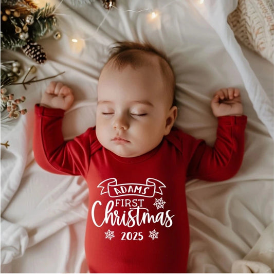 Baby wearing a red 'Adams First Christmas 2025' onesie lying on a white blanket with Christmas decorations.