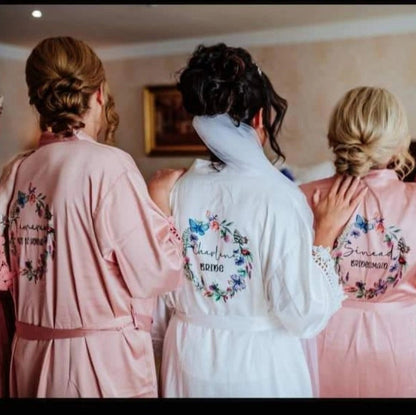 Bride and bridesmaids wearing personalised satin robes with floral wreath designs on the back, featuring names and wedding roles, printed by HanaLee Studios in Ireland.