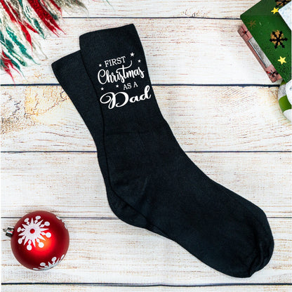Black socks with 'First Christmas as a Dad' text on a wooden surface with a red ornament.