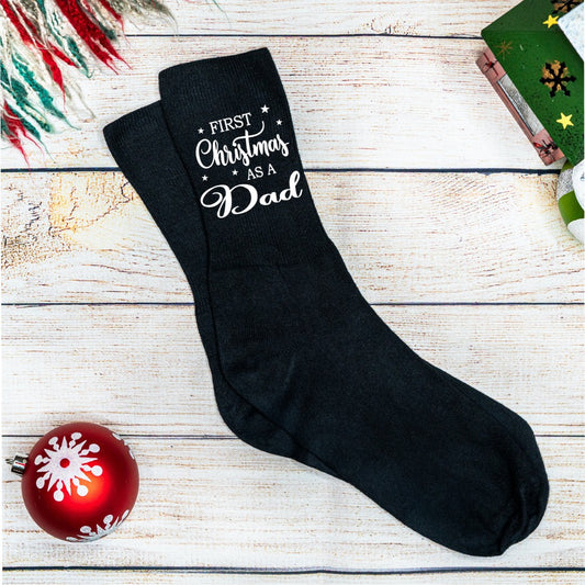 Black socks with 'First Christmas as a Dad' text on a wooden surface with a red ornament.