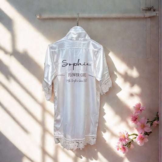 Personalised satin flowergirl robe in white with lace trim, customised with name ‘Sophie’ and wedding date, handmade in Ireland by HanaLee Studios.