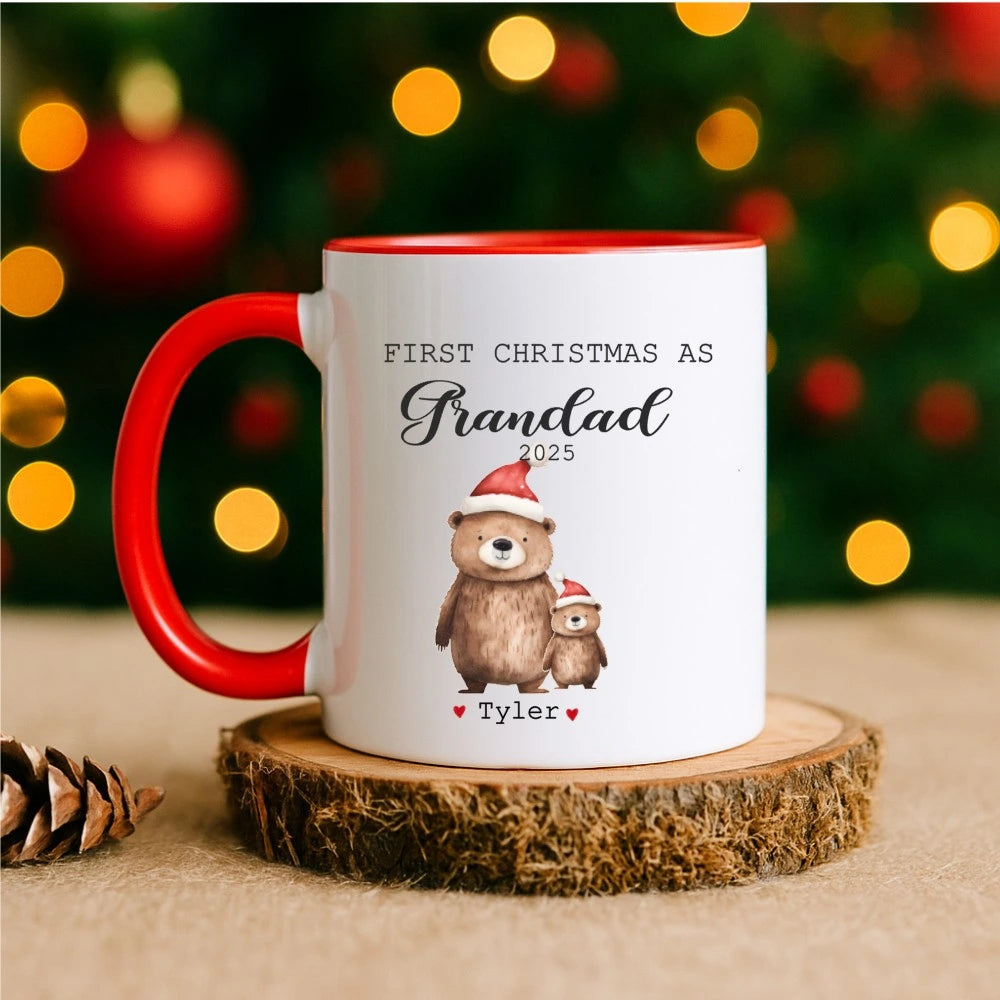 Mug with bear illustration and text 'First Christmas as Grandad 2025' on a wooden coaster with festive lights in the background.
