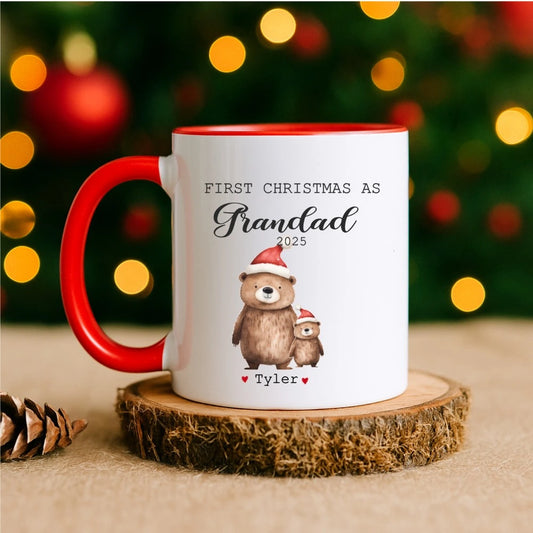 Mug with bear illustration and text 'First Christmas as Grandad 2025' on a wooden coaster with festive lights in the background.