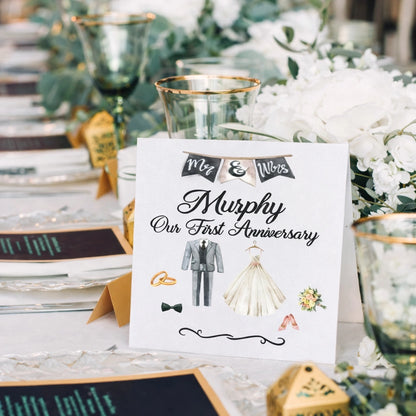 Personalised “Our First Anniversary” wedding card standing on an elegant reception table with green glassware, white floral centrepiece and gold details.