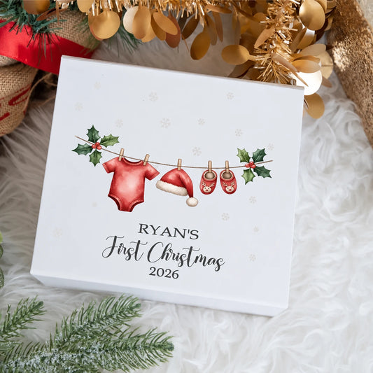 White personalised Baby's First Christmas gift box featuring baby's name, the year 2026 and a Christmas clothesline design with a red baby vest, Santa hat, reindeer booties and holly.