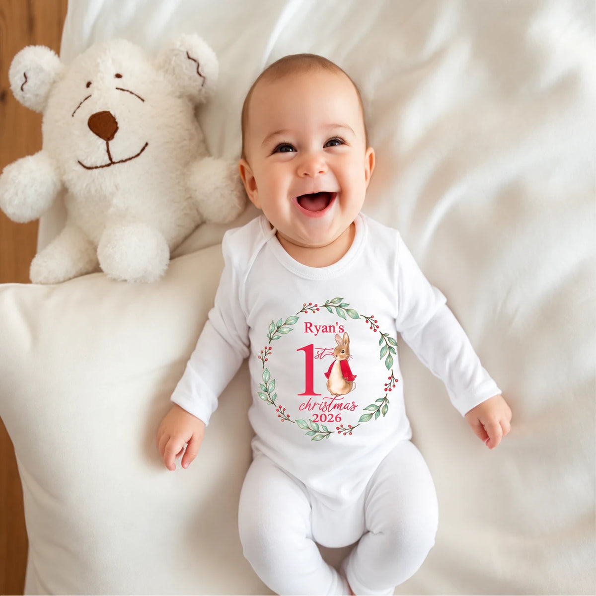 Smiling baby wearing a personalised white First Christmas 2026 babygro featuring a watercolour bunny in a red coat, surrounded by a green foliage wreath and red berries, beside a soft teddy bear.