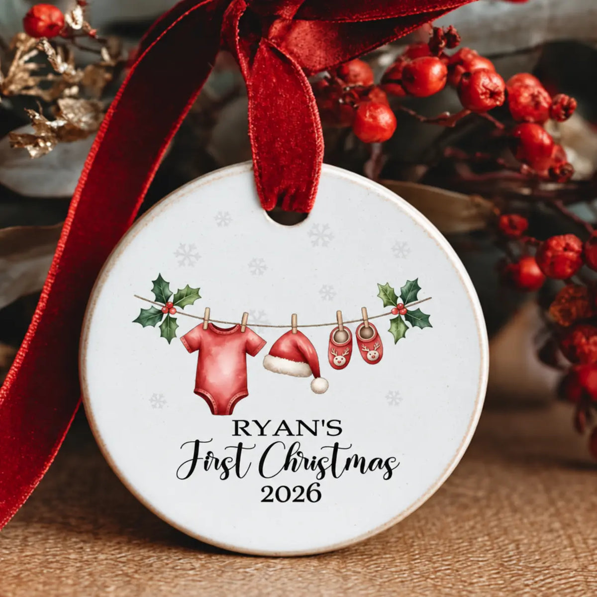 Personalised Baby’s First Christmas 2026 ceramic decoration featuring the child's name, a red baby vest, Santa hat and reindeer booties hanging on a Christmas clothesline, finished with a red velvet ribbon.