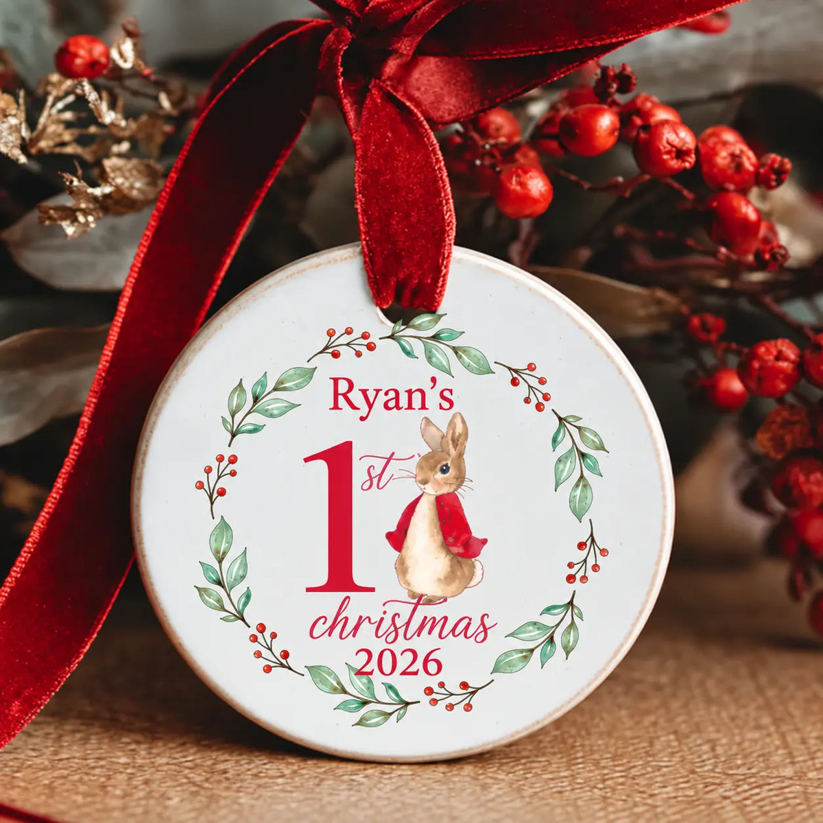Personalised Baby’s First Christmas 2026 ceramic decoration featuring a watercolour bunny in a red jacket, the child's name, a green foliage wreath with red berries and a red velvet ribbon.