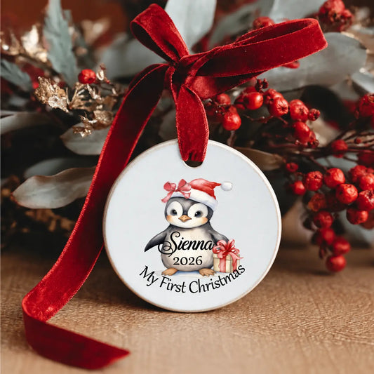 Personalised Baby’s First Christmas 2026 ceramic decoration featuring a cute baby penguin in a Santa hat, the child's name , a Christmas present and a red velvet ribbon.