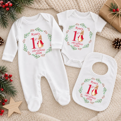 Personalised Baby's First Christmas outfit bundle featuring a white babygro, vest and bib with matching bunny, holly wreath and 2026 design, laid out on a neutral Christmas background.