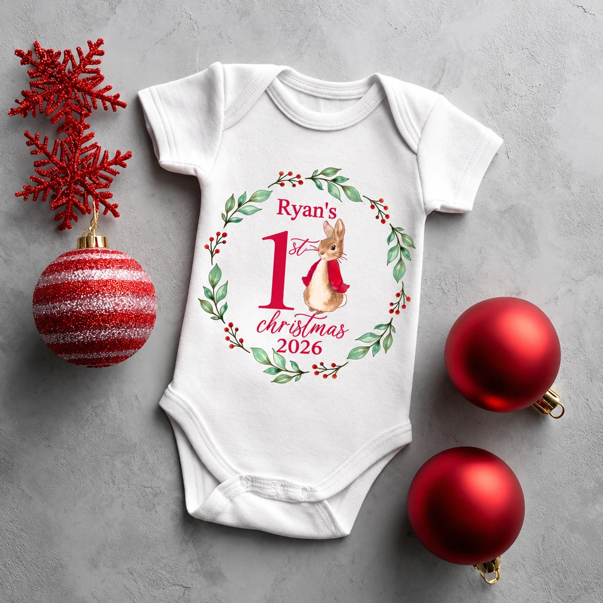 Personalised Baby's First Christmas 2026 white short-sleeved baby vest featuring a watercolour bunny in a red coat, surrounded by a green foliage wreath and red berries.