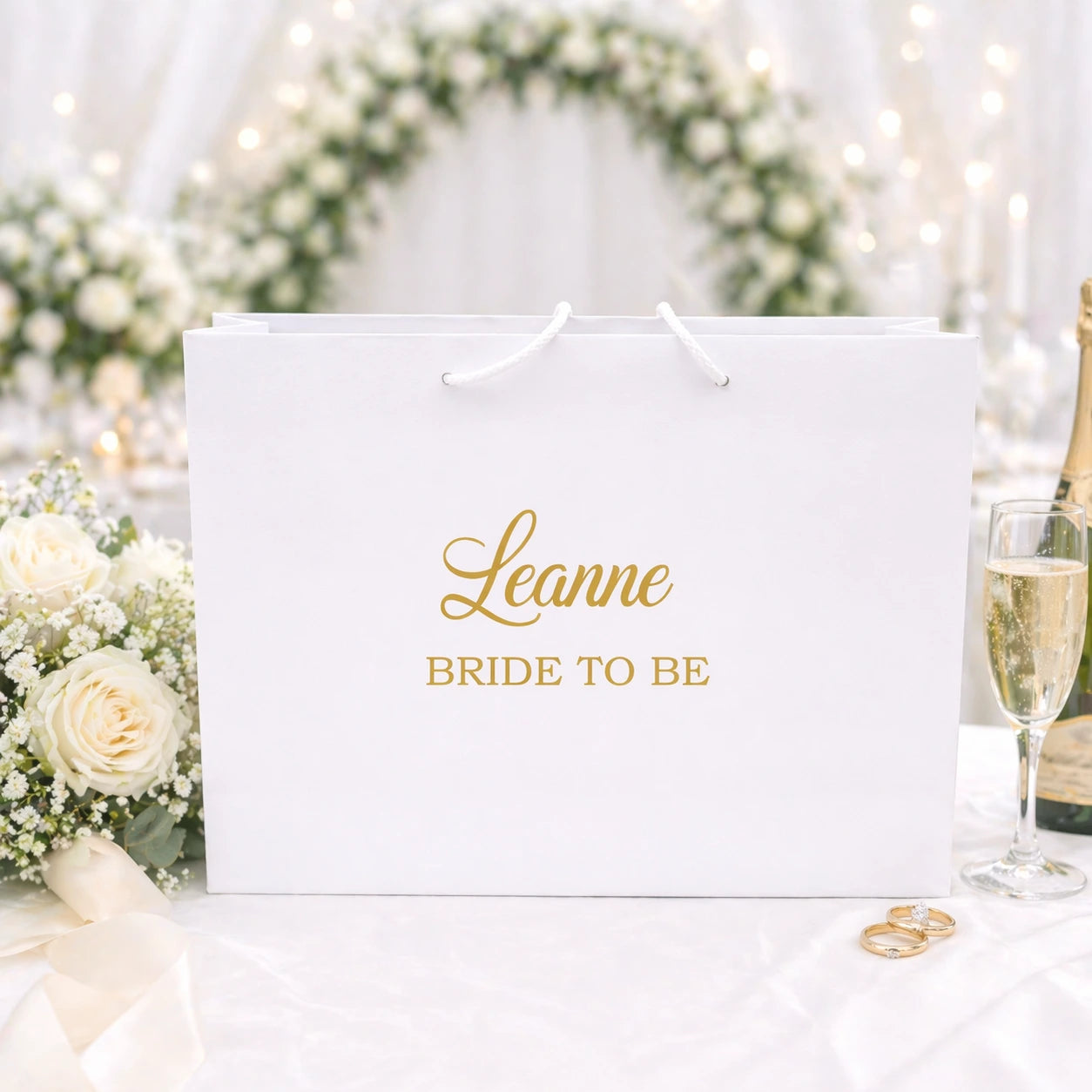 White personalised bride to be gift bag with gold name, styled with wedding flowers, champagne and rings in a soft bridal background.