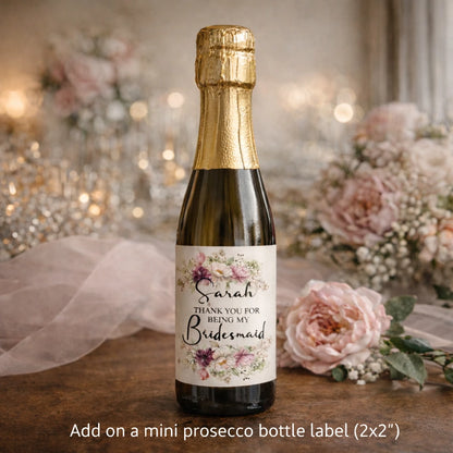 Mini bottle of prosecco with personalised floral bridesmaid label saying “Sarah – Thank You for Being My Bridesmaid” displayed on a table with soft pink wedding flowers.
