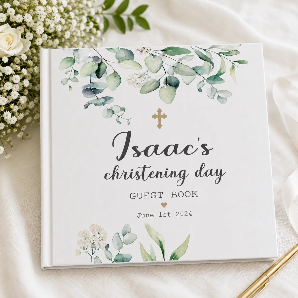 Personalised white christening guest book for Isaac with green eucalyptus design, gold cross and date, styled with white flowers and gold pen.