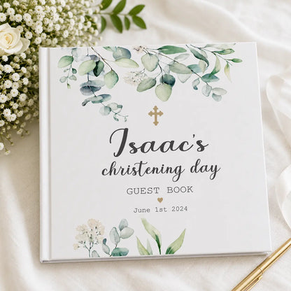 Personalised white christening guest book for Isaac with green eucalyptus design, gold cross and date, styled with white flowers and gold pen.