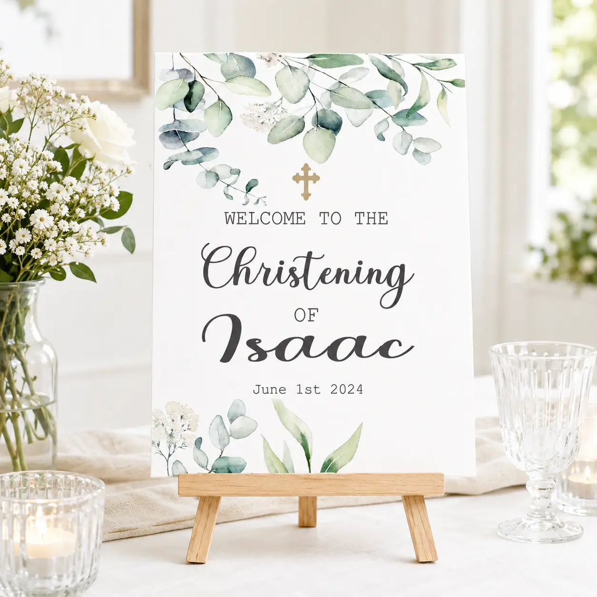 Personalised A3 christening welcome sign with green eucalyptus design and gold cross, displayed on a small wooden tabletop easel.