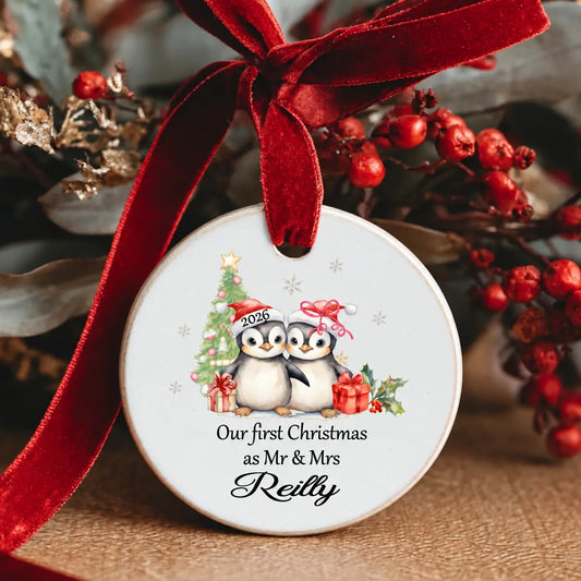 Personalised Our First Christmas as Mr & Mrs 2026 ceramic Christmas decoration featuring two cute penguins in Santa hats, a Christmas tree, presents and the surname, finished with a red velvet ribbon.