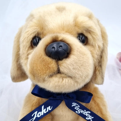 Golden plush puppy gift for a page boy, personalised with a navy satin name ribbon and displayed against a bright wedding-inspired background.