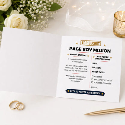 Open square white Page Boy Mission proposal card displayed on a white wedding-themed table with roses, candlelight and wedding rings. The inside right page features a mission briefing, page boy proposal message, wedding date and location fields, and mission acceptance checkboxes.