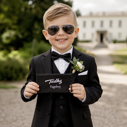 Young page boy wearing personalised aviator-style sunglasses and holding a custom black sunglasses case with his name printed on it, standing outdoors at a wedding venue in a formal black suit.