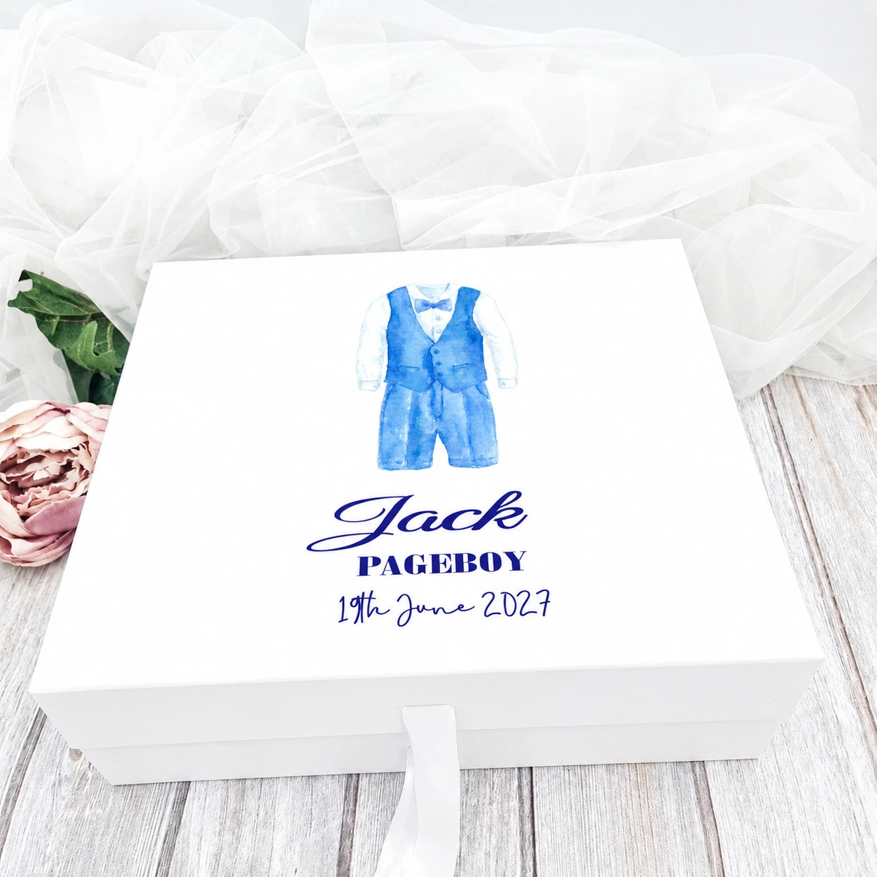 White personalised pageboy keepsake box featuring a blue watercolour pageboy suit design, custom name and wedding date, displayed on a wooden table with soft white wedding tulle and floral decorations in the background.