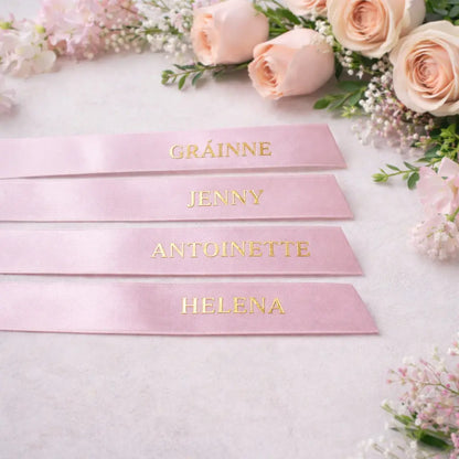 Four pale pink satin wedding ribbons personalised with metallic gold names, styled with blush pink flowers on a neutral background, ideal for bridal party and wedding styling