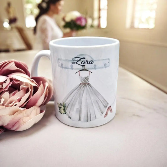 Personalised bridal mug featuring a wedding dress illustration with the name “Zara” above it, placed on a marble surface beside pink flowers, with a bride blurred in the background holding a bouquet.