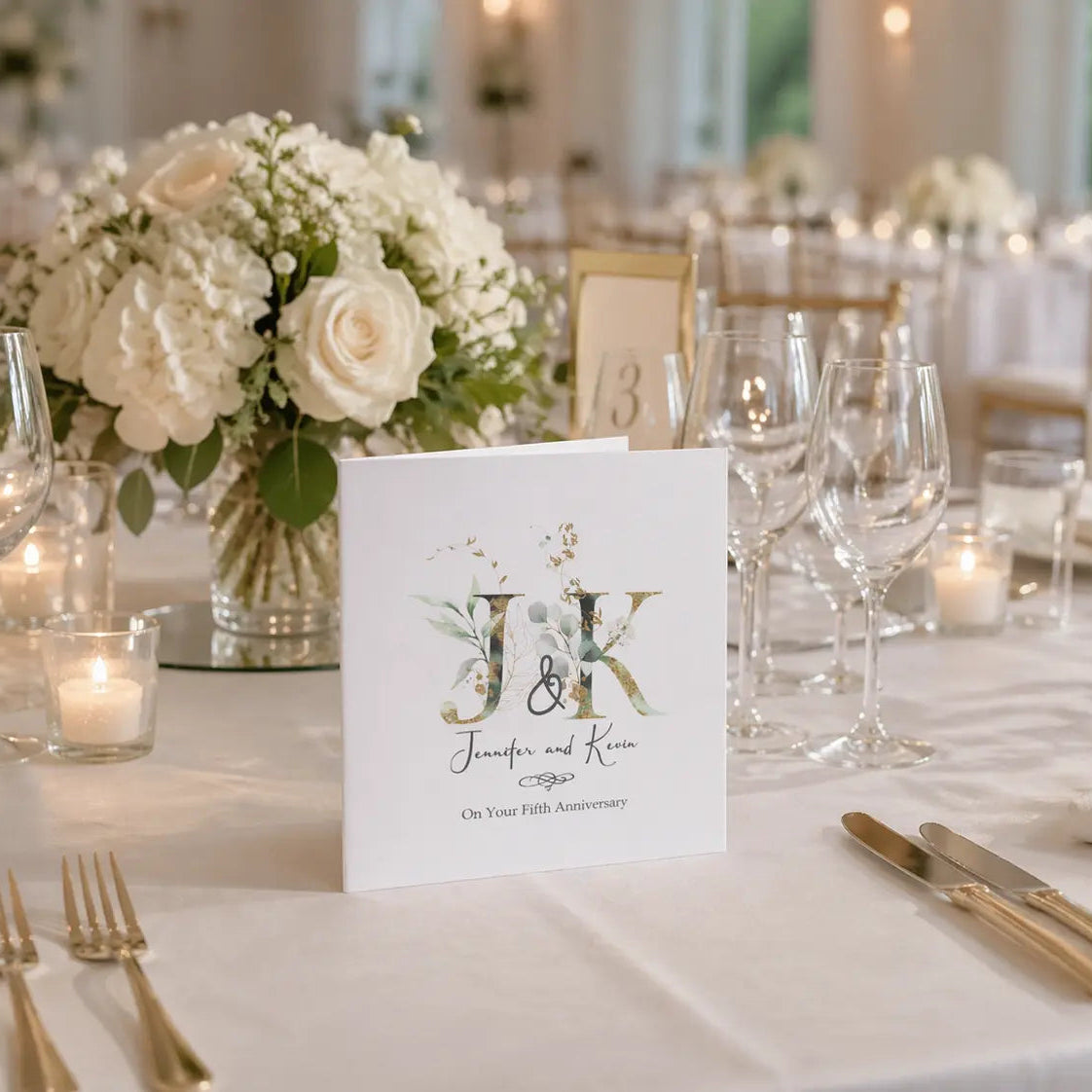 Personalised anniversary card with botanical monogram design displayed on an elegant wedding reception table with white flowers, candles and gold cutlery